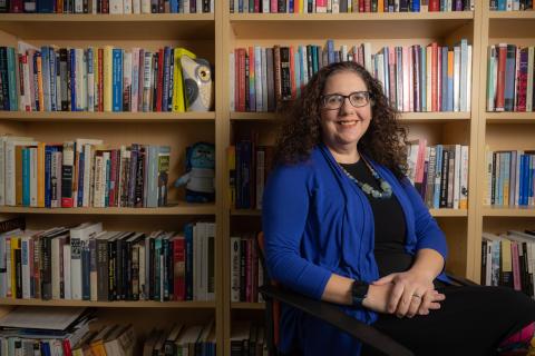 A white woman with curly brown sits in front of bookshelves